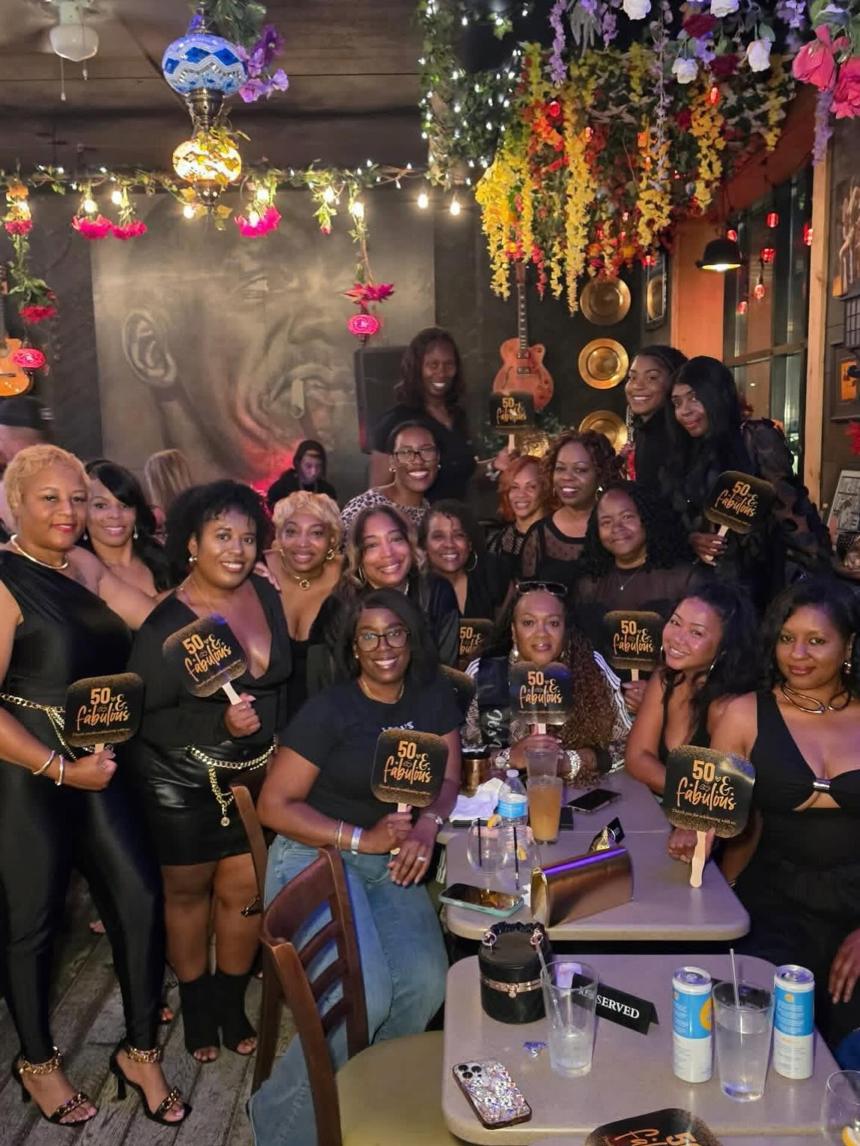 A large group celebrating together at Gibraltar under hanging flowers, string lights and a mosaic lantern, with guitars, gold plates and a painted mural on the wall behind them.