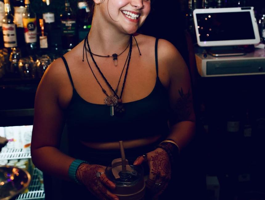 A smiling Gibraltar bartender with henna-painted hands passing a finished drink across the bar, backlit by bottles and warm lights.